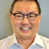 A smiling middle-aged man with glasses is wearing a white shirt. The background is a soft gray, creating a neutral and professional atmosphere.