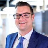 A smiling man in a blue suit and tie stands in a brightly lit office space. He has short brown hair and wears glasses, exuding a professional and approachable demeanor.