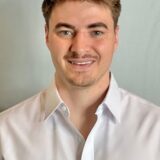 A young man with light brown hair smiles, wearing a white button-up shirt against a soft gray background.