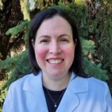 A smiling woman wearing a light blue lab coat stands outdoors, with green trees in the background.