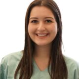 A smiling young woman wearing a light green medical scrubs top stands against a plain white background. Her long hair is down, and she appears friendly and approachable.