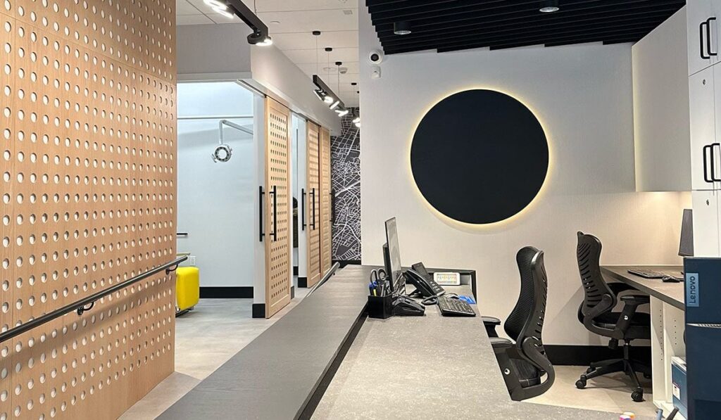 A modern office space featuring a sleek reception area with a long counter, two ergonomic chairs, and geometric design elements. The background includes private meeting rooms with wooden paneling and a circular design feature on the wall.