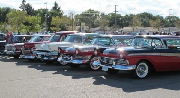 A row of classic retro cars in various colors parked side by side at an outdoor event, surrounded by trees and a blue sky.