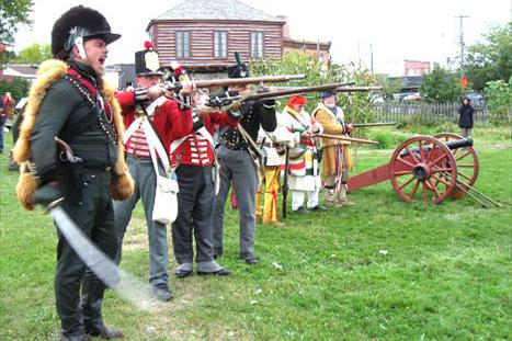 A group of historical reenactors in traditional military uniforms stands in a grassy field, preparing to fire their muskets. One soldier brandishes a sword, while a cannon is positioned nearby. A rustic building is visible in the background.