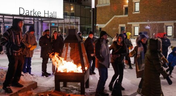 A group of people gathers around a fire pit outside Darke Hall, enjoying the winter evening. They are dressed warmly and appear to be engaged in conversation and laughter amidst the snow-covered ground.