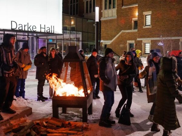 A group of people gathers around a fire pit outside Darke Hall, enjoying the winter evening. They are dressed warmly and appear to be engaged in conversation and laughter amidst the snow-covered ground.