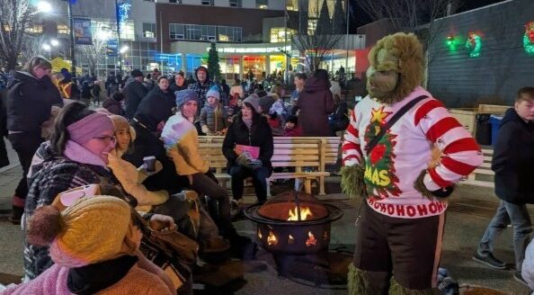 A crowd gathers around a fire at night, enjoying a festive atmosphere. In the foreground, a person dressed as the Grinch in a holiday-themed sweater interacts with attendees, while others are seated on benches, wrapped up in winter clothing.