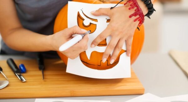 A person is using a stencil to carve a spooky face onto a pumpkin. They hold the stencil in place with one hand while using a marker with the other. Various carving tools and additional stencil designs are visible on a wooden surface nearby.