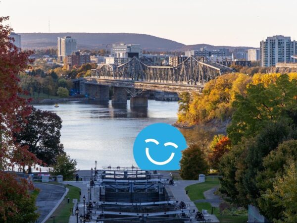 A scenic view of a river with a bridge in the background, surrounded by trees displaying autumn colors and city buildings on the horizon. A blue smiley face is overlayed on the image.