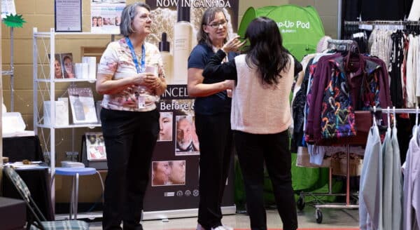 Three women are engaged in conversation at a trade show, surrounded by beauty products and clothing displays. The setting features promotional materials in the background.