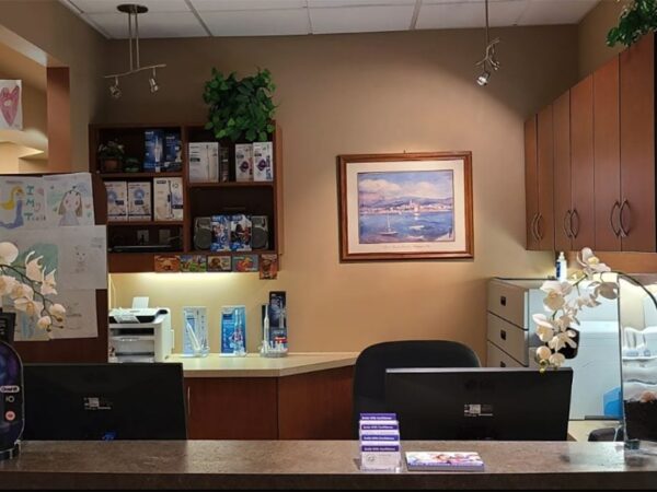 Reception area of a dental office with a desk, two monitors, decorative orchid plants, informational pamphlets, and wall art.