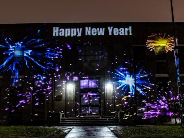 A building is illuminated with colorful projections of fireworks, with the words "Happy New Year!" displayed prominently at the top. The scene is set at night, adding a festive atmosphere to the celebration.