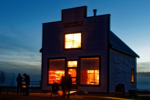 A small, illuminated house stands against a twilight sky, with warm light glowing from its windows. Silhouettes of people gather outside, creating a cozy and inviting atmosphere.