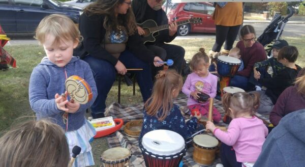 Children are engaged in a music activity outdoors, playing various percussion instruments while adults accompany them with a guitar. A sense of community and joy is evident as they explore rhythm together.