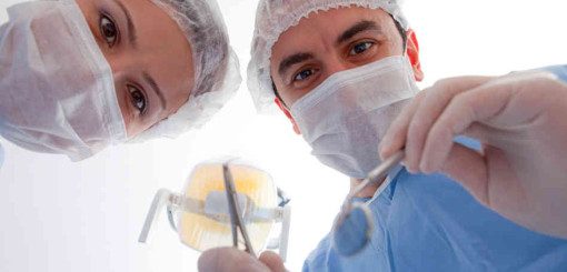 Two dental professionals in scrubs and masks are looking down towards the camera, holding dental tools. The background is bright, suggesting a clinical setting focused on dental care.
