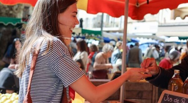 A smiling woman engages with a vendor at a bustling market under bright red umbrellas. She is exchanging money for goods, surrounded by colorful stalls and shoppers enjoying the lively atmosphere.