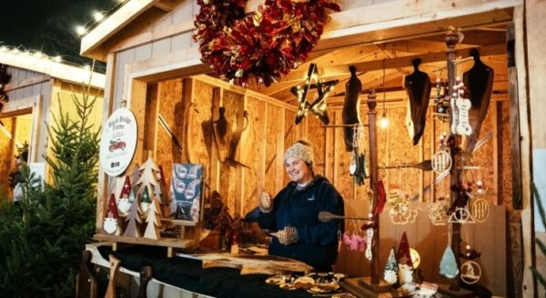 A cheerful vendor stands behind a wooden stall adorned with handmade crafts and decorations. A festive wreath hangs above, and various ornaments are displayed on the table, capturing the spirit of a holiday market. Warm lights illuminate the cozy atmosphere.