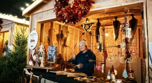 A vendor stands behind a wooden stall adorned with handcrafted ornaments and decorations, including wooden figurines and a wreath. The setting is festive, with warm lighting highlighting the charming display.
