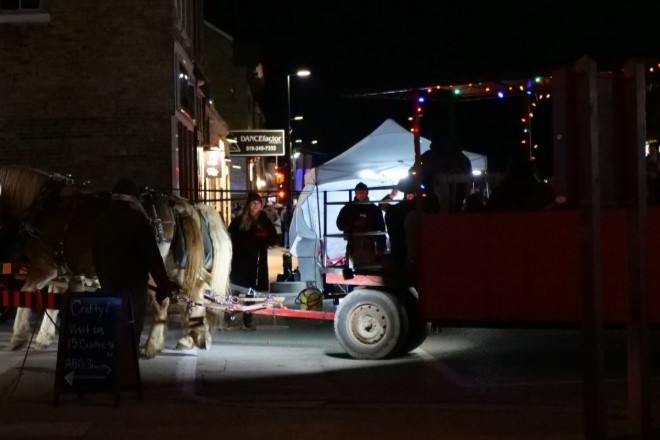A horse-drawn cart is parked at night, illuminated by festive lights. People are nearby, engaging with the scene, while a white tent stands in the background, hinting at a festive or community event.