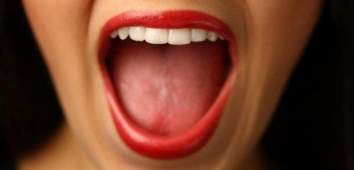 Gros plan d'une bouche ouverte avec du rouge à lèvres rouge vif, mettant en valeur des dents blanches et une langue visible. L'image capture une expression d'excitation ou de surprise.