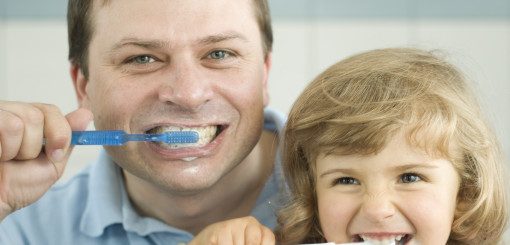 A smiling father and daughter brush their teeth together, both holding blue toothbrushes. The playful scene captures a moment of family bonding and encourages dental hygiene.