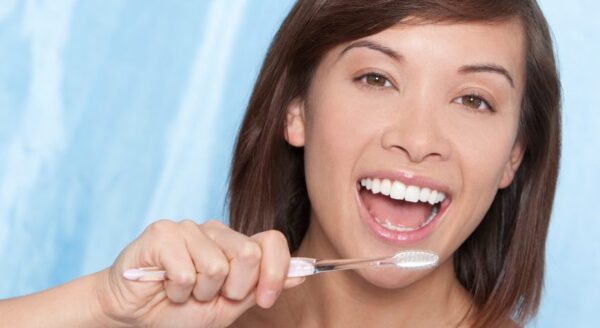 A smiling woman with medium brown hair brushes her teeth with a toothbrush, showing her bright smile. The background is light blue.