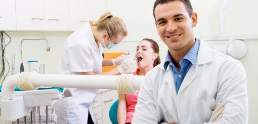Un dentiste en blouse blanche sourit avec assurance tandis qu'une assistante dentaire s'occupe d'un patient sur un fauteuil dentaire, qui semble subir un examen de contrôle. La clinique est lumineuse et bien équipée, ce qui témoigne d'un environnement dentaire professionnel.