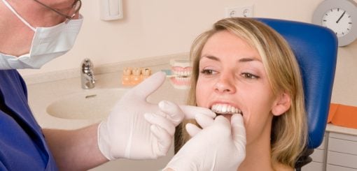 A dentist in gloves examines a woman's teeth while she sits in a dental chair, smiling slightly. The setting is a dental office with equipment visible in the background.