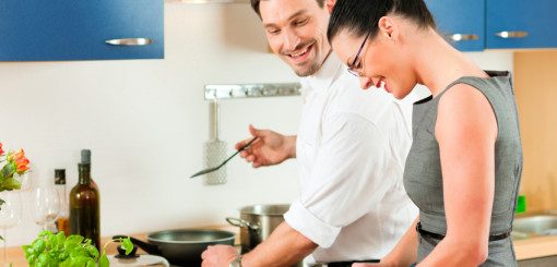 A man and a woman are cooking together in a modern kitchen, smiling and enjoying the experience. The man is stirring a pan, while the woman is cutting vegetables, creating a collaborative and friendly atmosphere.