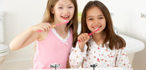 Two young girls are smiling and brushing their teeth by a bathroom sink. One girl is wearing a pink tank top, and the other is in a pajama top with a playful pattern. The scene conveys a cheerful and routine moment in personal care.