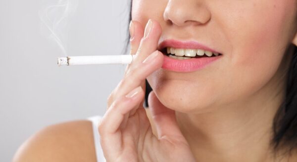 A close-up of a woman's face, smiling and holding a cigarette between her fingers. Wisps of smoke rise from the cigarette, and she has lightly colored lips and a relaxed expression.