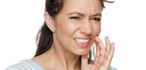 A woman with medium-length hair is grimacing and holding her jaw, expressing discomfort or pain, possibly from a toothache. She wears a light blue shirt and is looking towards the camera.