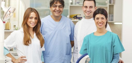 A group of four dental professionals, including two women and two men, stand together in a dental clinic. They wear scrubs and lab coats, smiling confidently at the camera, ready to provide care.