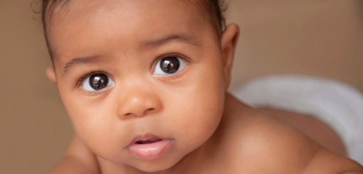 A close-up of a curious baby with large, expressive eyes and a neutral expression, looking directly at the camera. The baby has light brown skin and short, curly hair. The background is soft and out of focus, emphasizing the infant's face.