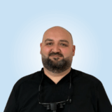 A smiling healthcare professional wearing a black shirt, with a beard and safety glasses around his neck, stands against a light blue background.