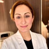 A professional woman wearing a white lab coat smiles confidently at the camera, standing in a modern medical environment with soft lighting.