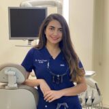 A smiling female dental professional in a blue scrub top stands beside a dental chair and equipment, with a monitor in the background, conveying a welcoming atmosphere in a dental clinic.