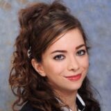 A young woman with curled brown hair and a bright smile, wearing a black jacket and a white shirt. The background features a soft, blue gradient.