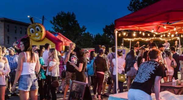 A lively night market scene featuring a bustling crowd enjoying food and festivities. Colorful lights illuminate vendor stalls, and a whimsical bee-shaped attraction is visible, enhancing the festive atmosphere.