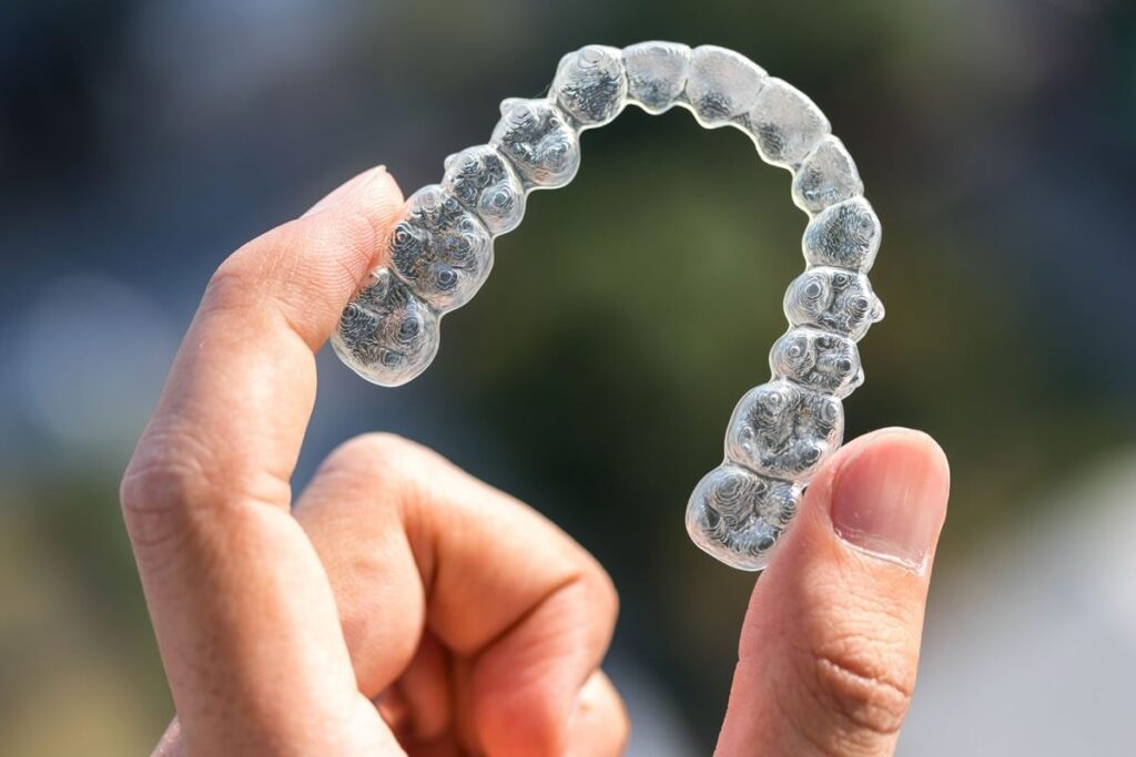 A person holds a clear plastic dental aligner in the shape of an arch, showcasing its transparent design. The background suggests an outdoor setting with blurred greenery.