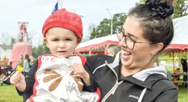 A woman joyfully holds a baby dressed in a red hat and a light-colored outfit, both smiling. They are outdoors during a festive event, with tents and greenery in the background.