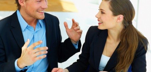 A man and a woman are engaged in an animated conversation, both wearing business attire. The man appears enthusiastic as he gestures while speaking, and the woman listens intently, smiling. The setting suggests a professional environment.