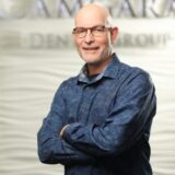 A smiling man with glasses stands confidently with his arms crossed, wearing a blue patterned shirt. The background features a softly textured wall with the words "Dental Group" prominently displayed.