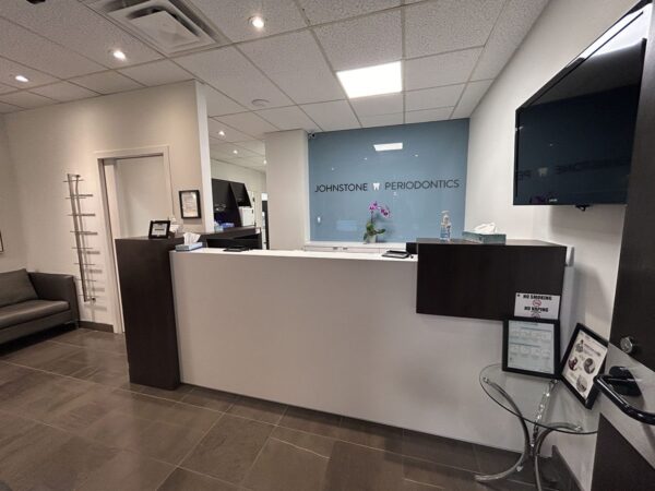 Interior of a medical reception area featuring a sleek white reception desk, a green accent wall with the clinic's name, an informational display, and a comfortable seating area. A television mounted on the wall and modern decor enhance the welcoming atmosphere.