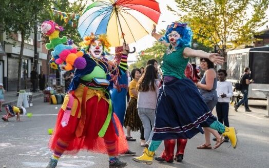 Two joyful clowns in colorful costumes dance on a lively street, one holding a vibrant umbrella. They are surrounded by a cheering crowd and green trees, embodying a festive atmosphere.