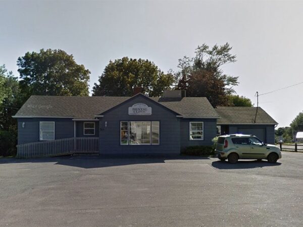 A blue building with a pitched roof, surrounded by trees, and a car parked in front. The scene is set on a clear day, highlighting a quiet area with a paved surface.
