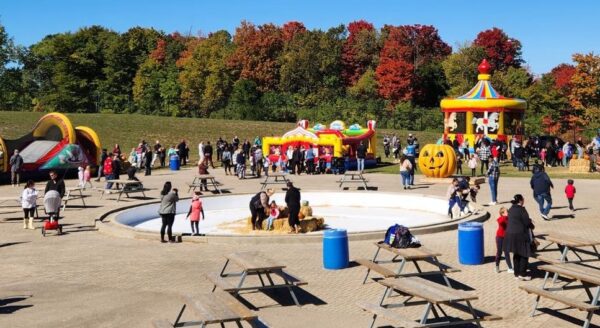A lively outdoor scene featuring families and children enjoying a fall festival. Attractions include colorful bounce houses, carnival games, and a carousel, set against a backdrop of autumn foliage. Picnic tables are scattered throughout the area.