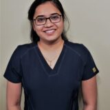 A smiling person wearing a dark scrubs top and glasses stands against a plain background, exuding a professional and approachable demeanor.
