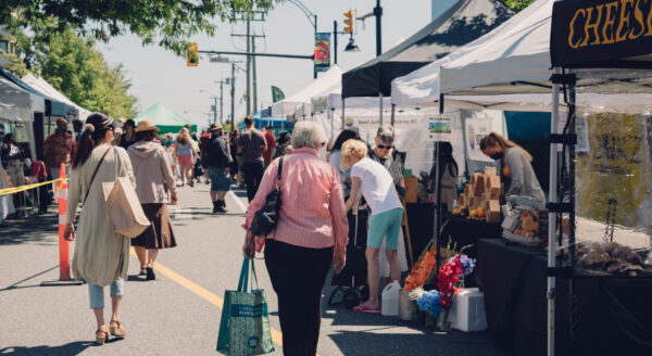A bustling outdoor market scene with various stalls featuring tents. People walk along the pathway, browsing products, with colorful displays of flowers and food items visible. Trees provide shade, creating a lively atmosphere filled with shoppers and vendors.