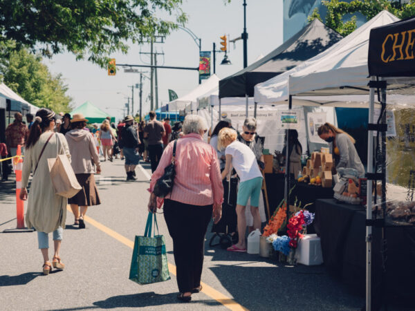A bustling outdoor market scene with various stalls featuring tents. People walk along the pathway, browsing products, with colorful displays of flowers and food items visible. Trees provide shade, creating a lively atmosphere filled with shoppers and vendors.